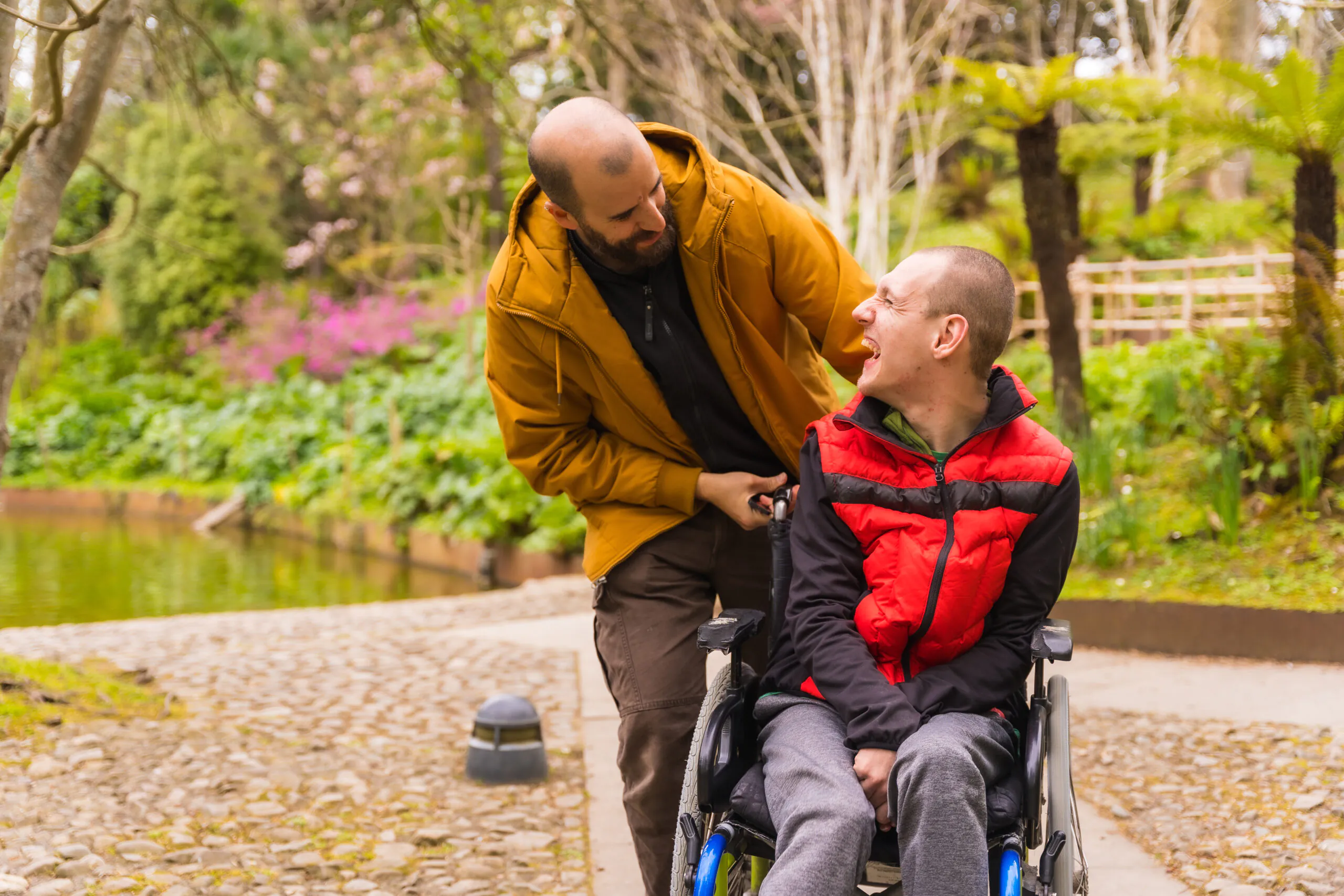 Young man in a wheelchair being pushed by a friend in a public city park, having fun in spring - positive behaviour support